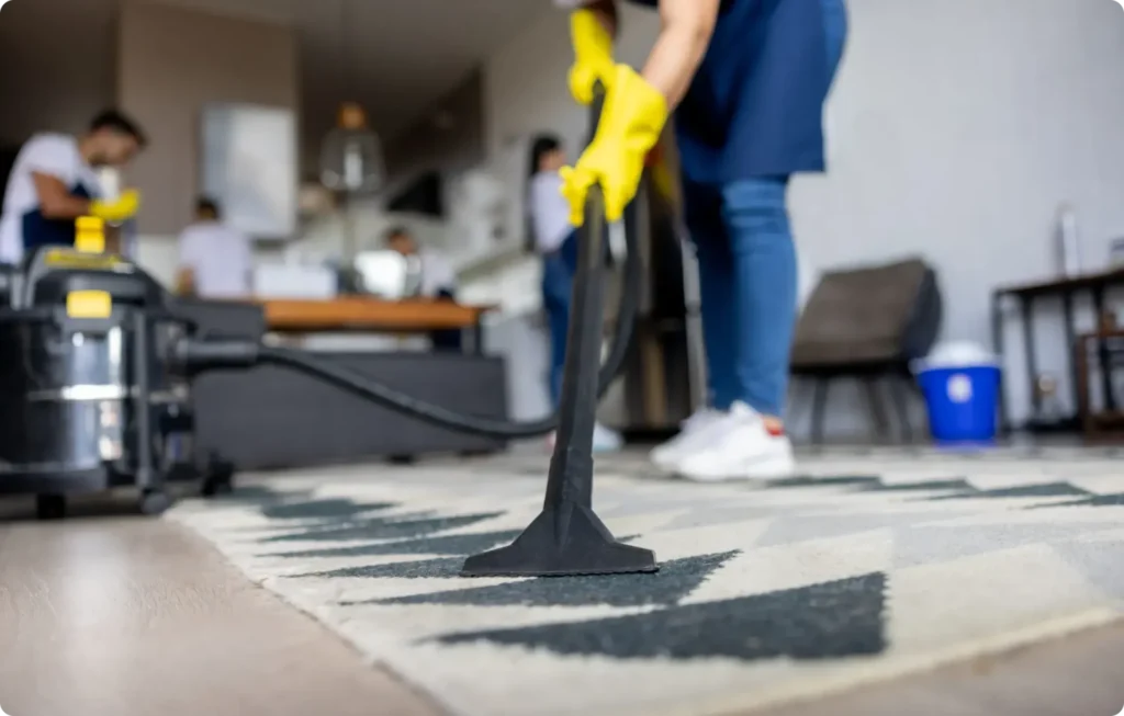 Close-up of a cleaner wearing a teal apron and yellow gloves, carrying a grey caddy filled with various cleaning supplies, brushes, and a green microfiber cloth. In the background, another person is mopping a shiny tiled floor.