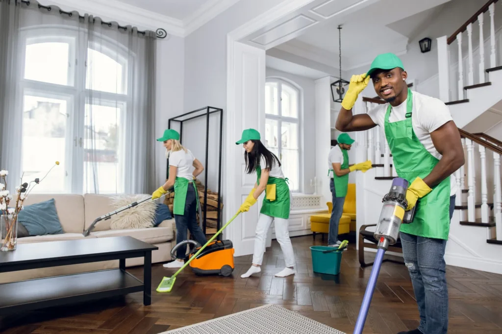 A professional cleaning team in green uniforms and yellow gloves offering maid cleaning services by cleaning a bright, modern living room with vacuums and mops.