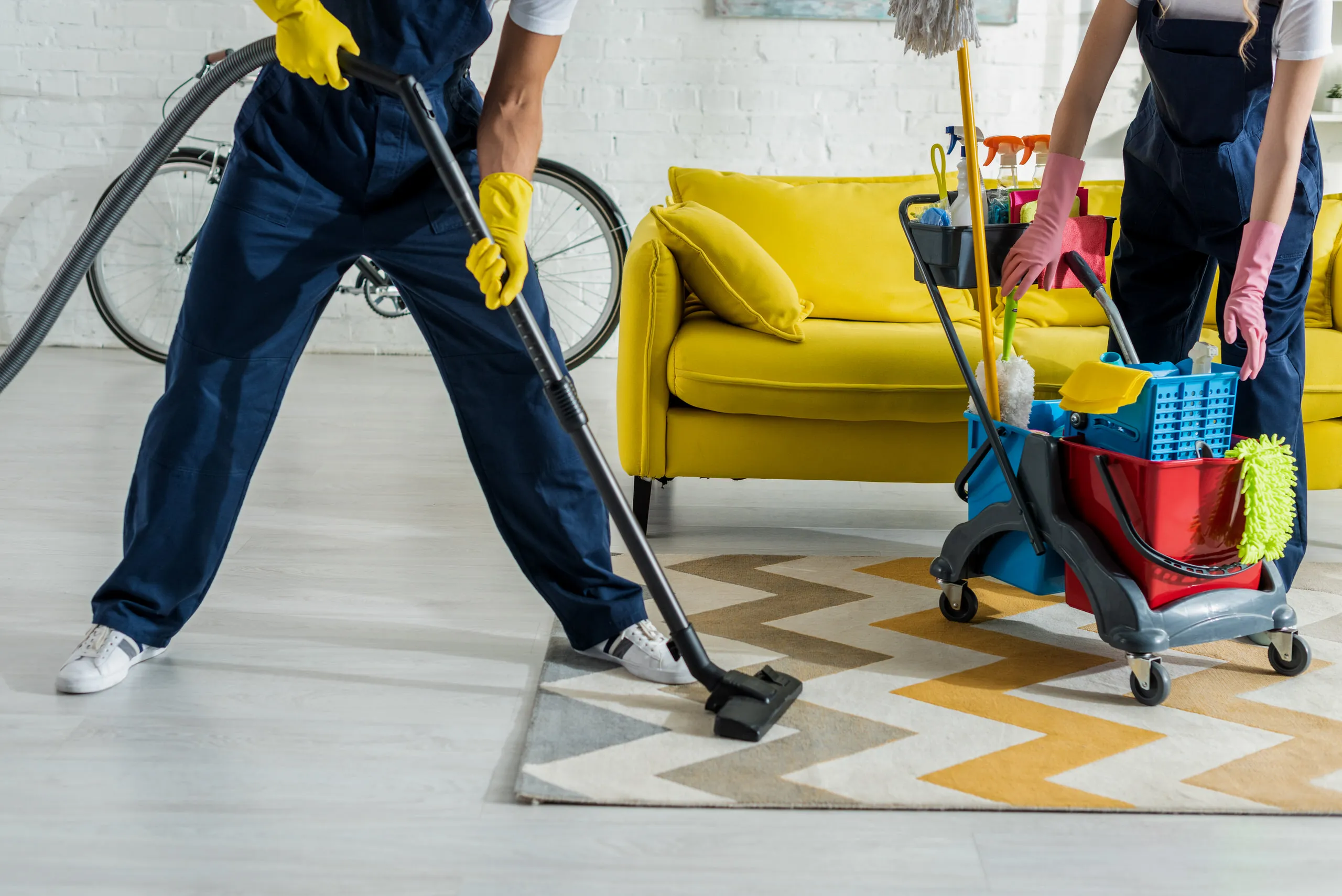 Two professional cleaners in dark blue overalls working in a living room. One person vacuums a grey and yellow chevron rug, while the other organizes a cleaning trolley stocked with sprays and brushes next to a bright yellow sofa.