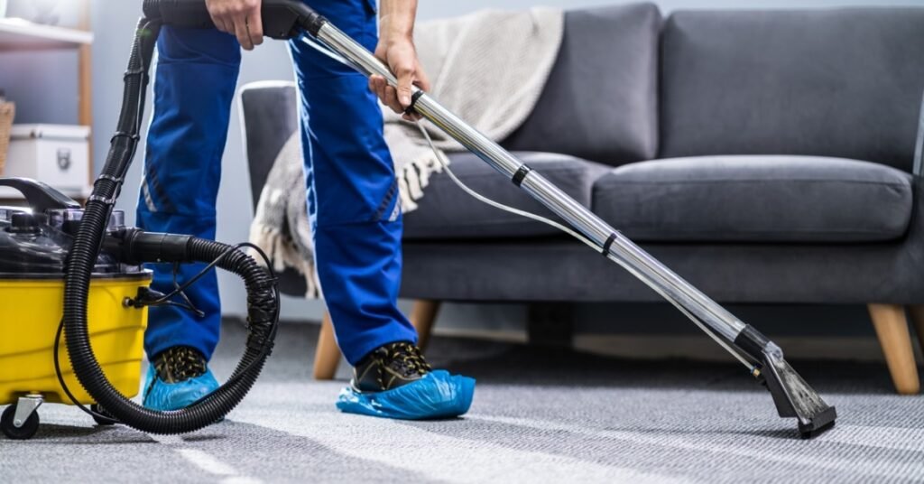 A person in blue overalls vacuums a carpet as part of regular cleaning maintenance.